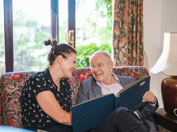 Besucherin und älterer Herr lachen zusammen auf dem Sofa und blättern in einer Mappe