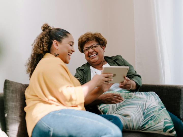 Jüngere und ältere Frau lachen gemeinsam auf dem Sofa über ein Tablet